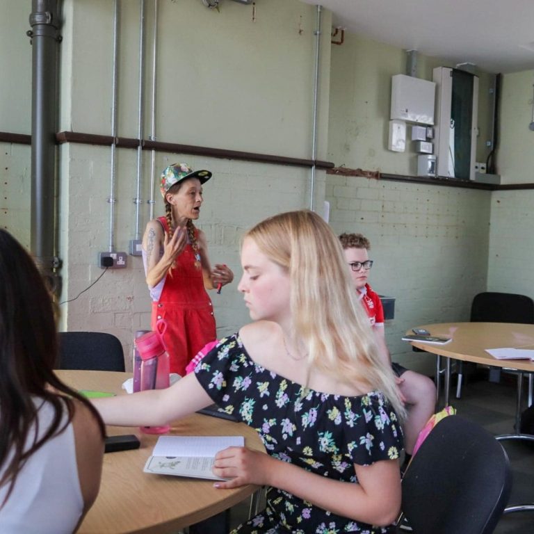 A lady in red dungarees speaks, while others engage at tables in a classroom setting.