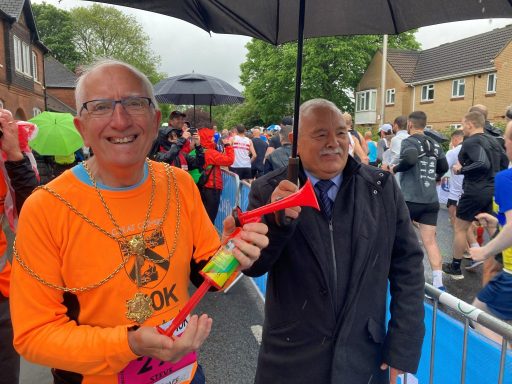 Two smiling men at an outdoor event, one holding a trumpet and the other an umbrella.