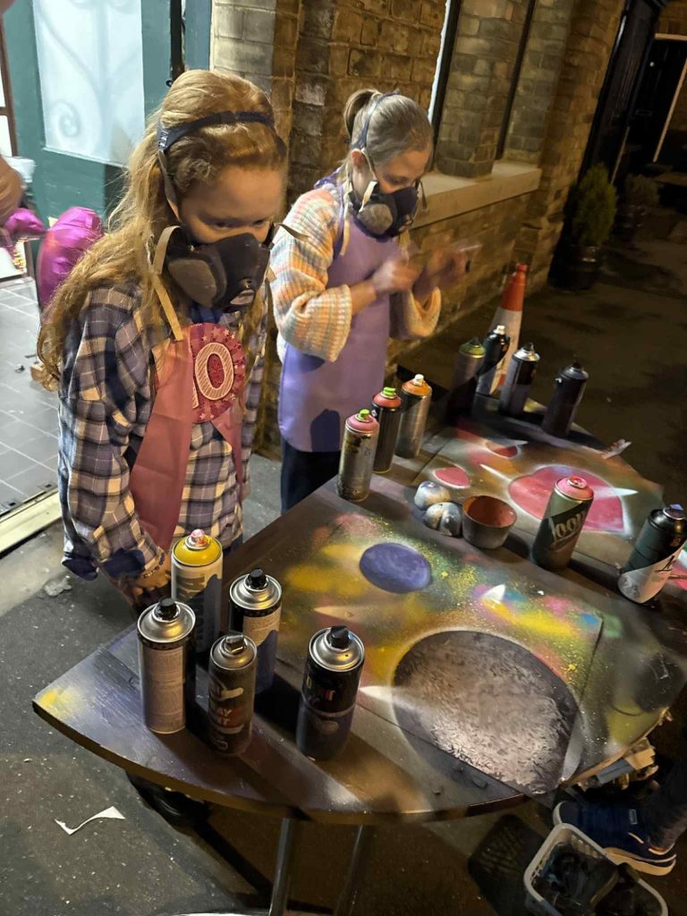 Two masked artists spray painting on a table outdoors, surrounded by art supplies.