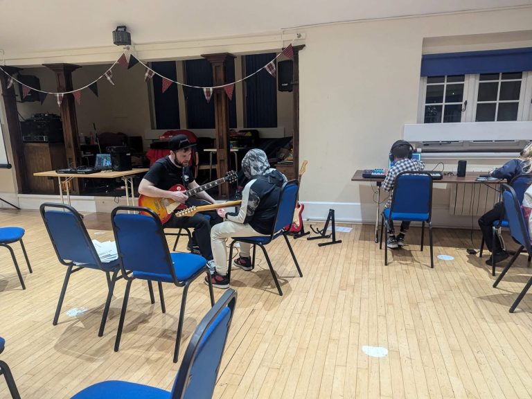 Two people play guitar in a room with chairs